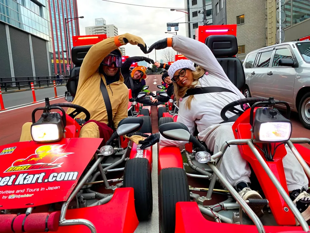 People in colorful costumes in red go-karts on an urban street, bending their arms to form a heart shape.