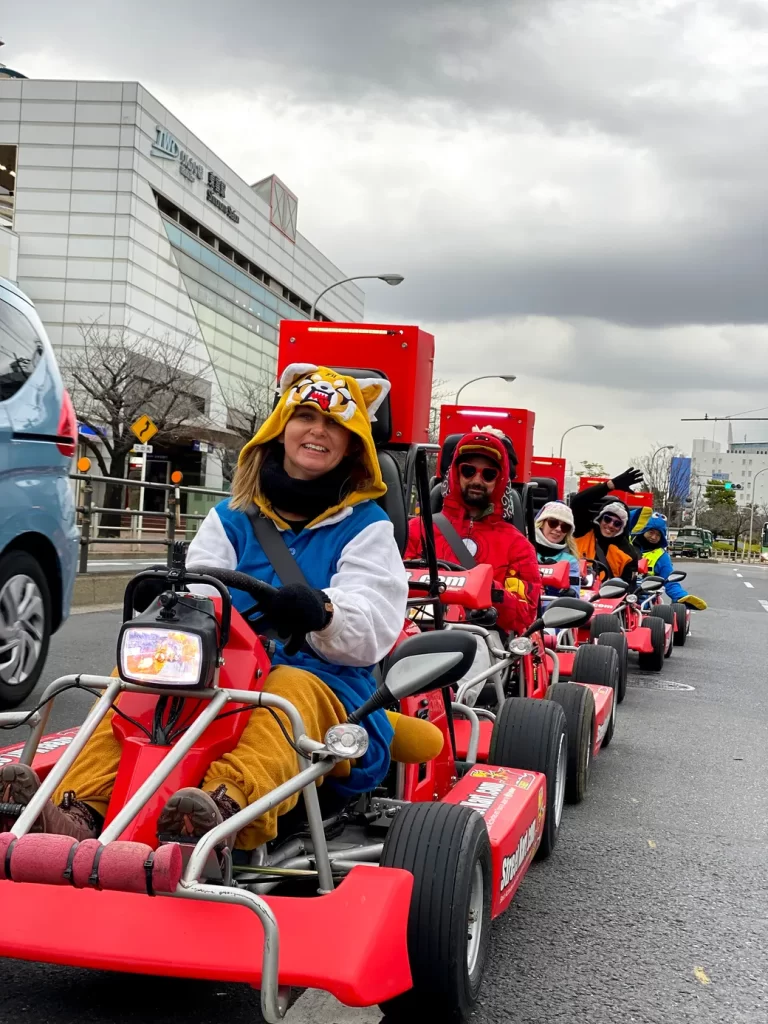 IMG_2602 - StreetKart Group of people riding red go-kart style parade carts on a city street, wearing colorful costumes and hats, smiling at the camera.