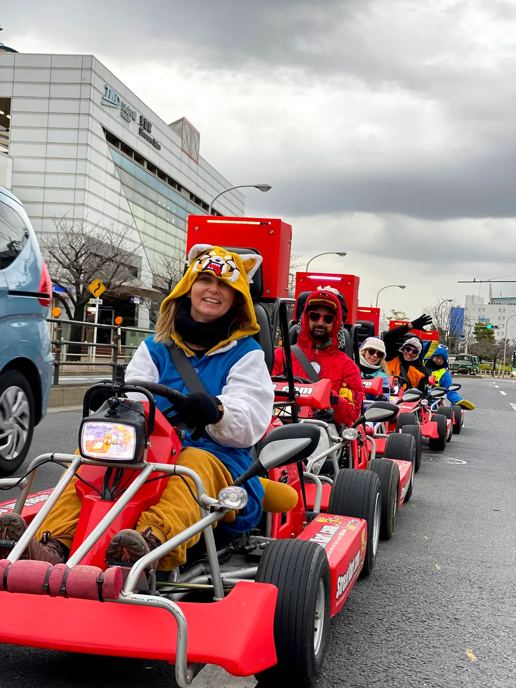 Group of people riding red go-kart style parade carts on a city street, wearing colorful costumes and hats, smiling at the camera.