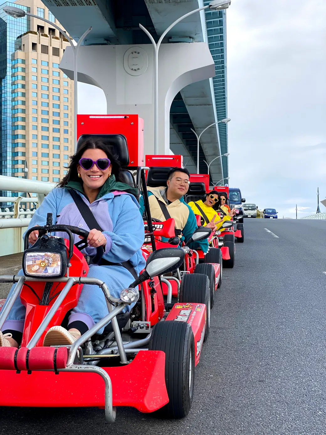 Group of people riding red go-karts on an elevated roadway, with a city skyline in the background under a bridge/overpass.